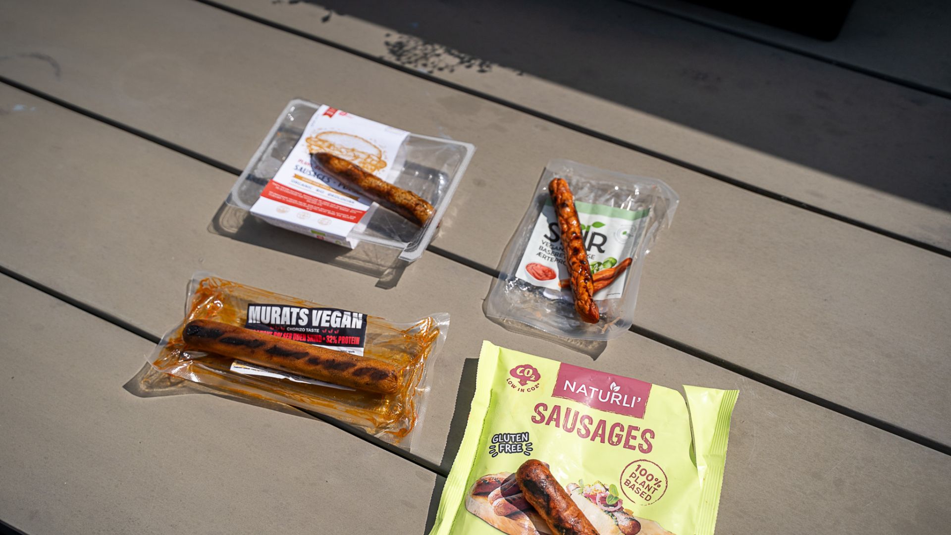 Fire pakker med veganske pølser ligger på bord i solen. Én pølse fra hver pakke er taget ud, så man kan se pølsen ordentligt.