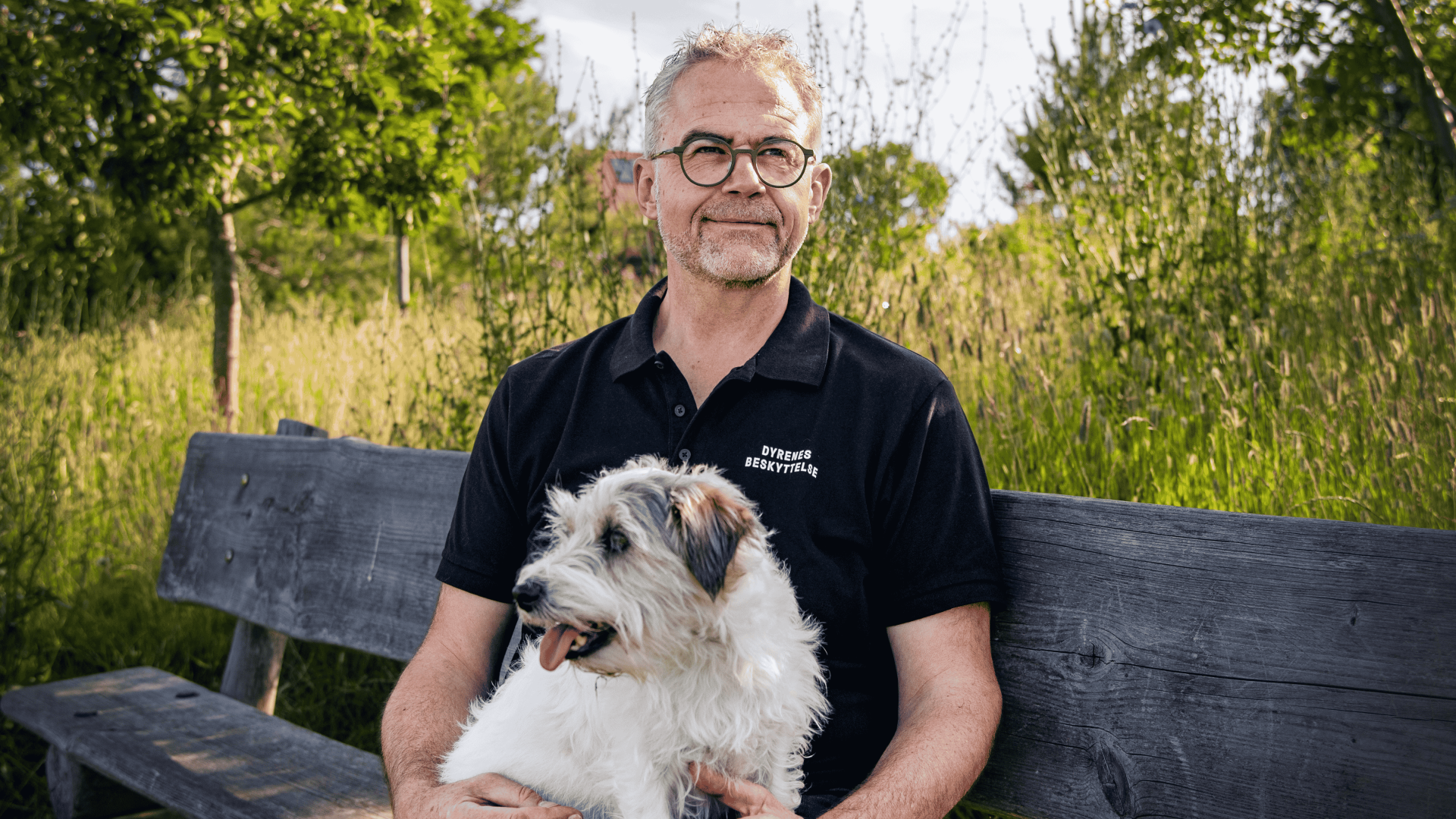 Jens Jokumsen, familiedyrschef i Dyrenes Beskyttelse, har haft med mange typer dyr at gøre igennem sit liv, men de sidste mange år har både hans privatliv og arbejdsliv været præget af familiedyrene. Foto: Benedicte Lorenzen / Dyrenes Beskyttelse. Jens Jokumsen sidder på en bænk i græsset med sin hund i skødet