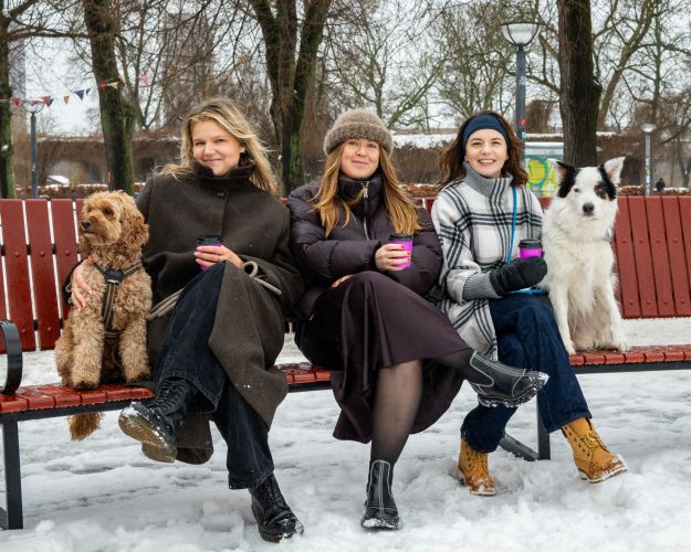 Tre unge kvinder sidder på en bænk i snevejr. Ved siden af sig har de to hunde, som også sidder på bænken. 