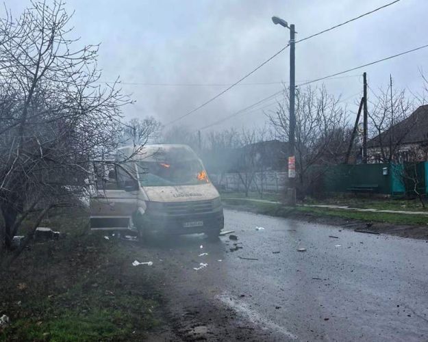 En varevogn er blevet ramt af en russisk drone i Ukraine, den holder på våd vej i gråt vejr. Luften er fuld af røg, og der er stadig flammer i bilens forrude.