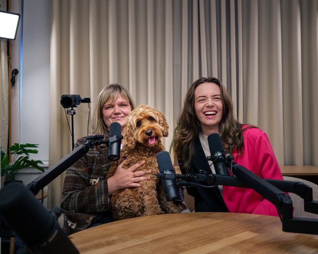 Værterne Katrine Nygaard og Nomi Forchhammer sidder i studiet med en krøllet hund.