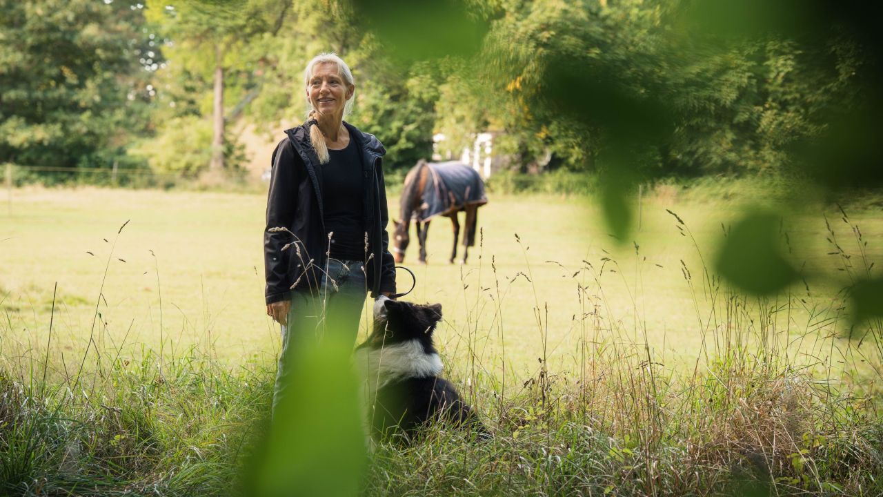 Maria Vang står i skovkanten foran en hestefold med sin hund i snor