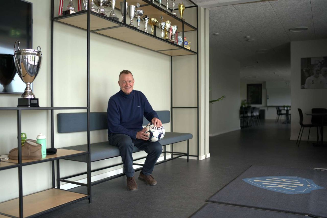 Per Frimann sidder på en bænk med en fodbold i hænderne og pokaler rundt om sig